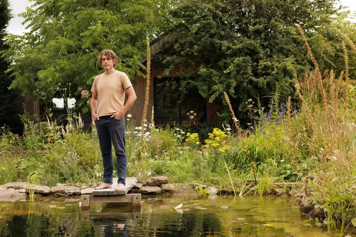 Will Tomson standing on a stone slab at the edge of a wildlife pond, planting and garden studio behind him