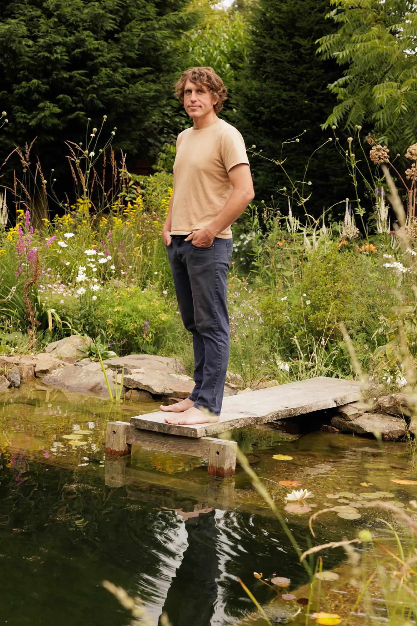 Will Tomson standing barefoot on a timber jetty over his natural swimming pond at Berrywell, wildflower planting behind — The Creative Gardener, 2026